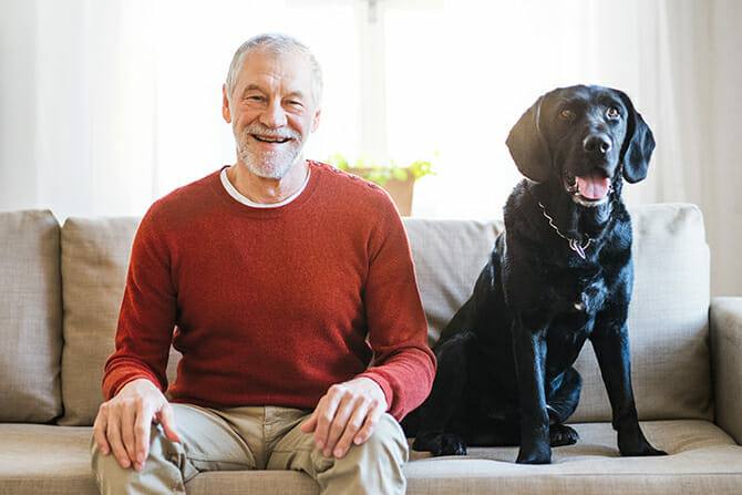 Therapiebegleithunde Trainer Ausbildung - älterer glücklicher Mann sitzt gemeinsam mit schwarzem großen Hund auf dem Sofa