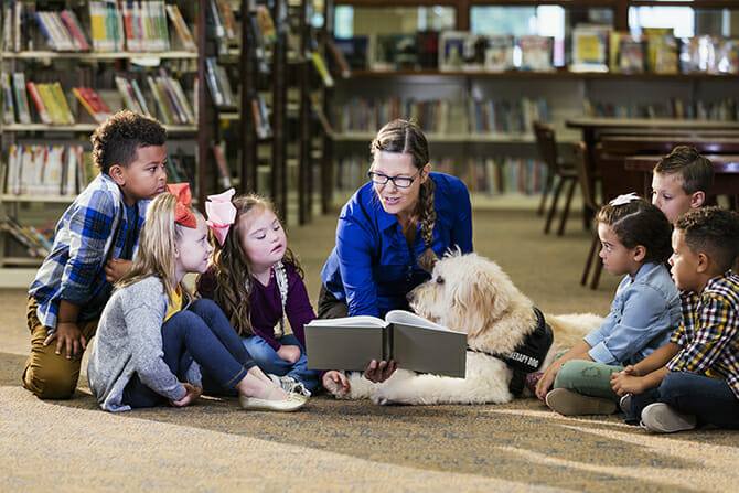 Therapiebegleithunde Trainer Ausbildung - Kinder in einer Bibliothek lesen gemeinsam mit einem Therapiebegleithund ein Buch
